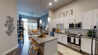 This well-appointed single-story residence showcases a thoughtfully designed open-concept layout that seamlessly connects the kitchen, dining, and living areas. The kitchen is a standout feature, boasting tall white shaker cabinetry that extends toward the elevated ceilings, granite countertops, a subway tile backsplash, and a full suite of stainless steel appliances including a gas range, microwave, dishwasher, and side-by-side refrigerator. A generously sized center island with granite counters and bar seating serves as both a functional workspace and a social hub. Pendant lighting above the island adds a refined touch.

The living room is anchored by a distinctive textured accent wall, ceiling fan, and a glass door leading to an outdoor area, filling the space with natural light. Wood-style plank flooring runs throughout the main living areas, adding warmth and continuity.

The bedrooms are carpeted and tranquil, each offering natural light through blinds-fitted windows. The primary bedroom connects to a walk-in closet with dual hanging rails and shelving. The bathroom features a white vanity with granite countertops and a glass-enclosed shower, maintaining the unit's clean, modern aesthetic throughout.