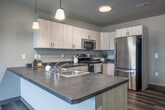 This single-floor unit features a clean, modern design with a neutral color palette throughout. The open-concept kitchen and living area serves as the heart of the space, where light wood-toned cabinetry pairs with dark countertops for a contemporary contrast. Stainless steel appliances, including a dishwasher and range, complement the functional layout, while pendant lighting and a ceiling fan add practical style. Wood-look plank flooring extends throughout the main living and kitchen areas, contributing warmth and visual continuity.

The bedroom areas are finished with plush wall-to-wall carpeting and feature large double-hung windows fitted with blinds, allowing for an abundance of natural light and elevated views of the surrounding streetscape and tree canopy. The neutral taupe walls and dark baseboards provide a grounded, cohesive aesthetic throughout these rooms.

The bathroom is well-appointed with a wide vanity featuring light wood cabinetry, ample drawer and cabinet storage, a large wall mirror with overhead lighting, and a white countertop sink. Wood-look tile flooring continues the unit's consistent design language. Overall, the space presents a bright, contemporary atmosphere with thoughtful finishes and a functional, open layout.