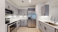 This single-floor residence showcases a clean, contemporary design with a cohesive aesthetic carried throughout every room. The kitchen is a standout feature, equipped with grey shaker-style cabinetry, matte black hardware, white quartz countertops, stainless steel appliances including a side-by-side refrigerator with ice and water dispenser, a built-in microwave, and a stainless range. A built-in wine rack above the refrigerator adds a refined touch, while light wood-look flooring ties the space together seamlessly.

The open-concept layout connects the kitchen to the living area via a peninsula with pendant lighting overhead, creating a natural flow for everyday living and entertaining. Dark, five-panel interior doors provide striking contrast against the bright white walls throughout the unit.

The bedrooms are bright and airy, each featuring ceiling fans, natural light from windows with blinds, and the same light wood-look flooring found throughout the home. The primary bathroom is generously proportioned, with grey cabinetry, quartz countertops, matte black hardware, and access to a walk-in closet. A secondary bathroom offers a full tub-shower combo with large-format white tile, a grey vanity, and quartz countertop. The overall ambiance is modern, bright, and move-in ready.