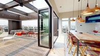 This contemporary rooftop amenity space offers a seamlessly connected indoor-outdoor experience across a single, well-appointed floor. The interior centers on a combined dining and kitchen area defined by striking design details, including a textured white-and-red geometric accent wall, stainless steel appliances, and a sleek white countertop. A long communal wood-top dining table surrounded by plaid-upholstered bar chairs serves as the focal point, illuminated overhead by a series of sculptural copper pendant lights that lend warmth to the modern aesthetic. Large windows and floor-to-ceiling glass sliding doors flood the interior with natural light while framing sweeping city skyline views. The kitchen is fully equipped with an induction range, range hood, under-counter refrigerator, microwave, and ample counter space. The interior flows directly onto a generous wood-deck terrace through oversized sliding glass doors. The outdoor living area is thoughtfully arranged with a sectional lounge, café-style seating, a built-in fire pit, and a dedicated outdoor kitchen complete with a gas grill and wall-mounted television. A pergola structure with string lights defines the covered portions of the terrace, creating an inviting atmosphere with unobstructed panoramic views of the surrounding cityscape.