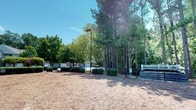 This outdoor dog park offers a well-maintained, open-air environment designed for pets and their owners to enjoy. The ground is covered with a generous layer of wood chips, providing a comfortable and low-maintenance surface for dogs to run and play. Mature pine trees and a variety of deciduous trees line the perimeter, offering ample natural shade on sunny days while contributing to a serene, wooded atmosphere.

A secure metal gate serves as the main entry point, ensuring pets remain safely within the enclosed space. Neatly trimmed hedges and ornamental shrubs add a touch of landscaping detail near the entrance, enhancing the overall appearance of the park. A standing light pole is positioned within the space, suggesting the area may be usable during early morning or evening hours.

A white bench near the gate provides a comfortable resting spot for pet owners to sit and supervise their animals. In the background, a brick building is partially visible, indicating the park is situated within a larger community or residential setting. The open canopy above allows for abundant natural light, creating a bright and welcoming outdoor space throughout the day.