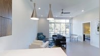 Located at 2304 Hillsborough St in Raleigh, NC, this modern single-floor space offers a bright and open layout designed with clean lines and contemporary finishes throughout. The main living and gathering area is defined by crisp white walls, large floor-to-ceiling windows that flood the interior with natural light and frame views of the surrounding greenery. Stylish pendant lighting in a brushed metallic finish hangs from the ceiling, complementing the recessed lighting and a modern ceiling fan that add to the room's functional elegance. The open-plan layout seamlessly connects a lounge area — furnished with a sleek dark sofa, blue accent chairs, and a patterned area rug — to a dining zone outfitted with a minimalist table and contemporary chairs. Light-toned tile flooring runs throughout, reinforcing the airy, modern aesthetic. The space strikes a balance between comfort and sophistication, making it well-suited for both relaxed gatherings and productive use. Its prime location on Hillsborough Street places it in the heart of one of Raleigh's most vibrant and accessible corridors.