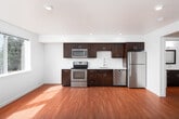 This single-floor unit presents a clean, modern design characterized by neutral white walls, recessed lighting, and warm hardwood-style flooring that extends through the main living areas. The open-concept layout connects the kitchen and living space seamlessly, creating a bright and functional environment. A large window in the main living area allows natural light to flood the room, offering views of surrounding greenery and providing a welcoming ambiance.

The kitchen is well-appointed with rich, dark-stained cabinetry offering generous upper and lower storage, white countertops, a stainless steel single-basin sink with a contemporary pull-down faucet, and a full suite of stainless steel appliances including a refrigerator, electric range, over-the-range microwave, and dishwasher.

The bedroom features neutral-toned carpet and crisp white walls, providing a quiet, comfortable retreat. The bathroom is finished in a clean, minimalist style with white walls, hardwood-style flooring, a white countertop vanity with dark cabinetry, and a full-size soaking tub with a shower and chrome fixtures. A hallway connects the rooms, maintaining a practical and efficient flow throughout the unit.