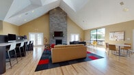 This club lounge is defined by its dramatic vaulted ceilings that rise to a striking peak, creating an open, airy atmosphere throughout the space. The centerpiece of the main lounge area is a floor-to-ceiling stacked stone fireplace, topped with a mounted flat-screen television and flanked by French doors that open to an outdoor deck, allowing abundant natural light to pour in. Comfortable leather seating is arranged around a patterned area rug in bold red, charcoal, and navy tones, complementing the warm hardwood-style flooring found throughout.

The lounge transitions seamlessly into a dining area featuring round tables and modern chairs, positioned near large windows with views of the outdoors. A dedicated computer workstation area offers high-top desks with desktop computers, illuminated by arched clerestory windows and fitted roller shades. A bold red accent wall bearing the "Club Lounge" branding adds energy to the space.

Additional features include a forage area with sleek cabinetry, a game zone with a foosball table, and metal railing details that define different zones within the open floor plan. The overall design balances casual comfort with functional amenities.