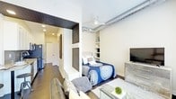 This single-floor studio space combines industrial and contemporary design elements to create a functional and stylish living environment. The open-plan layout integrates the sleeping and living areas seamlessly, maximizing the available space. Dark hardwood flooring runs throughout, grounding the neutral beige walls and creating a cohesive aesthetic.

An exposed metallic ductwork detail runs along the upper wall, nodding to an industrial design sensibility, while a ceiling fan with integrated lighting ensures comfort and practicality. Built-in wall shelving near the sleeping area provides storage and display space, adding both utility and visual interest.

A partial wall with bold, textured dark wallcovering serves as a design accent, visually separating the entryway corridor from the main living space. This entry area features a built-in bench with wall hooks, offering a convenient drop zone. The corridor leads toward what appears to be a kitchen and bathroom beyond.

The sleeping area accommodates a full-sized bed, while the living area features a sofa and glass coffee table, making efficient use of the compact footprint. A flat-screen television mounted on a dresser completes the functional arrangement of this thoughtfully designed space.