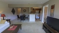 This single-story residence showcases an open-concept floor plan that seamlessly integrates the living, dining, and kitchen areas into one cohesive space. The living area is carpeted and anchored by a low-profile coffee table, providing a comfortable and understated setting. Adjacent to it, the dining area accommodates a round table with dark upholstered chairs, complemented by decorative wall art including a classic round clock, lending character to the space.

The kitchen is positioned at the rear and features a white peninsula that serves as both a workspace and a visual divider between the kitchen and dining areas. Cabinetry in warm wood tones lines the upper walls, while white appliances—including a range and refrigerator—are neatly integrated beneath an overhead ventilation hood. The kitchen floor transitions to tile, distinguishing it from the carpeted living and dining zones.

Natural light filters in from what appears to be a sliding glass door or large window on the right side of the space, brightening the interior. The overall ambiance is practical and welcoming, with neutral tones throughout creating a calm, cohesive aesthetic.