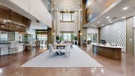 Located at 20050 N Cave Creek Rd in Phoenix, AZ, this sophisticated single-floor clubhouse offers a generously proportioned community space defined by elegant architectural design and high-end finishes throughout. The grand entrance lobby immediately impresses with its soaring double-height ceilings, a dramatic sputnik chandelier, floor-to-ceiling decorative columns, and large-scale artwork adorning the walls. Polished tile flooring reflects abundant natural light, while a central seating arrangement with a round table and upholstered chairs anchors the space. A staffed reception desk with fluted wood detailing adds a professional touch. Adjacent lounge areas feature plush neutral sofas, decorative plants, and tall windows offering views of the surrounding landscaping. The full-service kitchen is a standout feature, highlighted by a striking slatted wood ceiling installation, a curved island with bar seating, green marble countertops, stainless steel appliances, and a built-in wine refrigerator. A dedicated business center provides workstations with computers, while a formal conference room offers a polished boardroom table, executive seating, and a wall-mounted display. An indoor-outdoor lounge connects seamlessly to an exterior patio through a large glass garage-style door, creating an inviting flow to outdoor amenities.