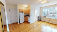 This single-floor unit is characterized by warm honey-toned hardwood floors that run throughout the main living areas, lending a cohesive and inviting feel to the space. The open layout connects the living area to the kitchen, which is tucked into a defined alcove. The kitchen is equipped with light oak cabinetry extending to the upper walls for ample storage, dark countertops, a tile backsplash, and white appliances including a gas range, refrigerator, and sink. A ceiling fan with a light fixture overhead serves the main living area, which also features a cast-iron radiator and a window-mounted air conditioning unit, providing year-round climate control. Natural light enters through windows that offer views of the surrounding neighborhood. The bedroom is a well-proportioned, bright room with its own ceiling fan, a window with exterior views, and the same polished hardwood flooring. The bathroom is finished in all-white, featuring tile flooring, a tub and shower combination with tiled surround, a pedestal-style vanity with mirrored medicine cabinet, and a window that provides natural ventilation and light.