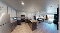This single-floor residence offers a thoughtfully designed open-concept layout that seamlessly connects the kitchen, dining, and living areas. The kitchen is a standout feature, showcasing dual-tone cabinetry with white upper cabinets and dark lower cabinets, warm wood-toned countertops, a mosaic tile backsplash, and a full suite of stainless steel appliances including a refrigerator, electric range with microwave above, and dishwasher. A peninsula with pendant lighting above provides additional prep space and casual dining potential. Light wood-look flooring flows throughout the main living areas, enhancing the bright, airy feel of the space. The living room is generously sized with room for a full seating arrangement, wall-mounted art display shelves, and a large sliding glass door that floods the space with natural light and offers outdoor views. The bedroom is a comfortable retreat, featuring a ceiling fan, a dedicated desk nook by the window, and ample wall space for artwork. The bathroom is finished with clean white subway tile surrounding a full soaking tub and shower combination with chrome fixtures. The entryway, accessible via a solid wood door, features light flooring and electrical panel access, providing a practical and welcoming transition into the home.