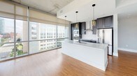 Located in the heart of downtown Durham at 511 S Mangum St, this contemporary urban residence offers a striking blend of industrial and modern design. The open-concept living area commands attention with soaring exposed concrete ceilings, rich hardwood flooring, and a dramatic wall of floor-to-ceiling windows that frame sweeping city views and open to a private balcony. The chef's kitchen is a standout, featuring dark navy cabinetry with gold hardware, a large white quartz waterfall island, subway tile backsplash, stainless steel appliances, and elegant pendant lighting. Bedrooms are finished with plush carpeting and large windows that continue the urban panorama, with exposed concrete columns adding architectural character. The primary bathroom is a sophisticated retreat, showcasing bold black penny-tile accent walls, a dual-sink navy vanity with gold hardware, white quartz countertops, and matte black fixtures. A second bathroom offers a clean white tiled tub-shower combination with matte black accents. Bedrooms include access to organized walk-in closets, completing this polished downtown living experience.