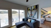 This contemporary single-floor space makes a strong design statement from the moment you step inside. The living area serves as the heart of the space, anchored by a sleek, dark sectional sofa adorned with patterned accent pillows in teal and yellow. A striking pair of large-scale abstract portrait paintings dominates one wall, infusing the room with vibrant color and artistic character that sets the tone for the entire space.

Natural light pours in through a generous sliding glass door, which opens onto a private outdoor balcony furnished with a wire accent chair — ideal for relaxing while taking in the surrounding landscape views. The clean white walls and smooth ceiling with a ceiling fan enhance the airy, open feel of the interior.

The overall ambiance is modern and curated, blending minimalist architecture with bold decorative choices. The combination of the expansive glazing, contemporary furnishings, and eye-catching artwork creates a space that feels both stylish and comfortable. The seamless indoor-outdoor connection further adds to the sense of openness, making this a thoughtfully designed space well-suited for both relaxing and entertaining.