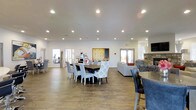 This single-floor community space is designed to foster social interaction and relaxation in an open, airy environment. The layout flows seamlessly from a central dining area to a comfortable lounge, creating a versatile setting for both casual gatherings and structured activities.

The dining area serves as the heart of the space, anchored by a large dark-wood table surrounded by an eclectic mix of upholstered and patterned chairs. Recessed lighting throughout the ceiling provides consistent, well-distributed illumination that enhances the warm tone of the hardwood-style flooring underfoot.

Adjacent to the dining area, the lounge features plush seating arrangements alongside a striking stone fireplace, offering a cozy retreat. A wall-mounted television and a large decorative board game display — reminiscent of a classic Scrabble board — add to the inviting, community-centered character of the space.

Natural light filters in through multiple glass-panel doors and windows along the perimeter, connecting the interior to the outdoors and contributing to the bright, open ambiance. A coffee and beverage station is conveniently positioned nearby, while a separate fitness area is visible through glass partitions, rounding out the amenities available within this thoughtfully designed communal facility.