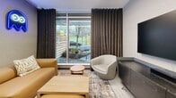Located at 137 Keith Rd E in North Vancouver, BC, this single-floor lounge space offers a thoughtfully designed living area that balances modern aesthetics with a relaxed, social atmosphere. Though compact in footprint, the room is efficiently arranged to maximize both comfort and functionality.

The living room serves as the central feature of the space, anchored by a large wall-mounted television positioned above a sleek, dark-toned media console. A soundbar and decorative greenery accent the console, adding warmth to the contemporary design. The walls are finished in textured grey, lending the space a sophisticated, neutral backdrop.

Seating includes a tan leather sofa adorned with patterned cushions, a rounded light-grey accent chair, and a low wooden coffee table — all arranged atop a layered area rug. A floor-to-ceiling window framed by long, dark drapes introduces natural light and provides views of the surrounding streetscape, enhancing the sense of openness.

Playful neon signs — including a "Movie Night" display and retro gaming icons — add a lively, casual character to the space. A large potted plant near the entrance contributes a natural element, while the light-toned carpet flooring ties the overall aesthetic together cohesively.