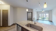 Located at 3655 Prairie Waters Dr in Grand Prairie, Texas, this single-floor residence offers a well-designed open-concept layout that seamlessly connects the kitchen and living areas. The kitchen is a standout feature, showcasing rich dark-stained cabinetry, a subway tile backsplash, stainless steel appliances including a microwave, range, and dishwasher, and a granite-topped island with an undermount sink. Pendant lighting and track lighting illuminate both the kitchen and adjoining living space, enhancing the modern aesthetic throughout. Light wood-look flooring extends across the main living areas, adding warmth and continuity to the open floor plan. The living area is bright and airy, benefiting from a large sliding glass door that opens to a private balcony, offering views of the surrounding neighborhood. A ceiling fan provides additional comfort in this space. The bathroom features a full soaking tub framed by large-format tile surround with a built-in niche, complemented by matching wood-look flooring. The layout also includes additional rooms accessible via interior doors, providing functional separation of spaces. Overall, the residence presents a clean, contemporary interior with quality finishes throughout.