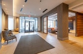 This modern building lobby offers a polished and well-appointed first impression. The main entrance features a sleek glass-and-steel door system with a vestibule that opens into a spacious, light-filled corridor finished with large-format tile flooring. Warm cove lighting runs along the ceiling, creating an inviting ambiance that complements the recessed can lights throughout. Textured accent columns and walls in muted taupe tones add architectural depth and visual interest to the space.

Off the main corridor, a dedicated lounge area is accessible through sliding shoji-style panel doors framed in rich wood trim. This room features a tray ceiling with pendant lighting, a bold teal accent wall, and comfortable seating with a sofa and armchairs, making it an ideal gathering space. Adjacent to this area, a sheer metallic curtain partition defines a flexible secondary space with tile flooring and additional seating.

A separate alcove houses a business or amenity station equipped with a wall-mounted display and a built-in console. The elevator bank is cleanly integrated into a dedicated alcove, featuring stainless steel doors with modern wall sconces on either side. Throughout the lobby, accent chairs in vibrant upholstery are thoughtfully placed, adding pops of color. The overall design balances functionality with contemporary elegance, making for a refined and welcoming communal environment.