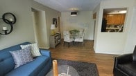This single-floor residence showcases an open-concept layout that connects the living and dining areas in a cohesive, functional arrangement. Rich hardwood flooring runs throughout, providing warmth and visual continuity across the space.

The living area accommodates comfortable seating, anchored by a plush area rug and a low-profile coffee table. A blue sofa with decorative accent pillows contributes a pop of color against the neutral, light-toned walls.

The dining area is positioned toward the rear of the open space and features a glass-top table surrounded by patterned upholstered chairs. A pendant light fixture with a dark drum shade hangs overhead, providing focused illumination. A decorative metal wall piece adds an artistic accent above the dining space, while a sideboard with a table lamp offers additional storage and ambient lighting.

A passthrough or opening to the right hints at an adjacent kitchen, featuring wooden cabinetry, creating an easy connection between cooking and living areas. A textured ceiling and clean white trim framing doorways and walls complete the well-appointed interior.