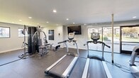Located at 9007 Ten Mile Rd in Knoxville, Tennessee, this well-equipped fitness center offers a comprehensive workout environment across a single floor. The space is thoughtfully designed with rubber flooring throughout, providing durability and safety for a wide range of exercise activities.

The main workout area is lined with True-brand cardio equipment, including multiple treadmills and an elliptical machine, all positioned to take advantage of large floor-to-ceiling windows and a glass door that floods the space with natural light and offers views of the outdoor surroundings. A wall-mounted flat-screen TV provides entertainment during workouts.

A dedicated strength training zone features a multi-station cable machine, complemented by a full dumbbell rack with a workout bench and a stability ball, offering a complete free weights experience. A separate, open stretching and functional training area provides ample room for floor exercises and is equipped with medicine balls and yoga mats.

Recessed lighting throughout ensures consistent illumination across all areas. A staircase connects the fitness center to adjacent building areas. The clean, neutral color palette of white and gray walls contributes to a bright, modern, and motivating atmosphere, making this facility a well-rounded amenity for residents of the community.