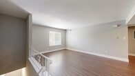 This two-bedroom, one-bathroom unit is thoughtfully designed to maximize its comfortable footprint across a single floor, accessed via an interior staircase with a decorative white railing. The open living area greets residents at the top of the stairs, featuring warm wood-look laminate flooring, soft neutral gray walls, and clean white baseboards that lend a cohesive, modern feel throughout. Natural light filters in through windows fitted with blinds, enhancing the bright and airy ambiance.

The kitchen is a standout feature, outfitted with white cabinetry, a teal subway tile backsplash, stainless steel appliances including a refrigerator and microwave, and a peninsula counter with a white surface that opens toward the main living space. An interior pass-through window connects the kitchen area to an adjacent space, adding architectural interest.

Both bedrooms are carpeted for added comfort and include windows with outdoor views. The primary bedroom is generously sized and benefits from a dedicated closet. The full bathroom is finished with large-format floor tiles, white subway wall tiles, and a tub-shower combination accented by a striking striped tile feature wall with modern chrome fixtures.