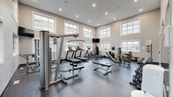 Located at 5501 Prefontaine Ave in Regina, Saskatchewan, this Fitness Suite is a thoughtfully designed exercise facility offering a full range of workout options within a bright and open single-floor layout. The space is defined by its impressively soaring double-height ceilings, which lend a sense of openness and airiness rarely found in residential fitness amenities. Large, multi-pane windows arranged on multiple walls — including an upper clerestory row — flood the interior with abundant natural light while offering views of the surrounding neighbourhood. The flooring consists of durable, dark rubber tile matting, ideal for supporting heavy equipment and high-impact activity. The fitness suite is comprehensively equipped with cardio machines including treadmills and an elliptical trainer, as well as a variety of strength training stations such as cable machines, a multi-function weight machine, adjustable benches, and a full dumbbell rack. A wall of mirrors along one side enhances the sense of space and allows users to monitor their form during exercise. A wall-mounted television provides entertainment, and a water dispenser is conveniently accessible. The neutral warm-grey walls and recessed lighting contribute to a clean, modern aesthetic that is both functional and inviting.