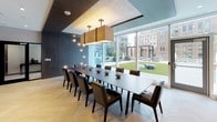 This elegantly designed single-floor space functions as a premium private dining and entertaining suite. At its centerpiece is a generously sized dark wood dining table surrounded by leather upholstered chairs, capable of accommodating a large group for formal gatherings or meetings. Overhead, striking square pendant lights with warm metallic finishes anchor the dining area, while a delicate cluster of globe lights adorns an adjacent wall, adding an artistic touch to the overall ambiance.

The most dramatic architectural feature is the expansive floor-to-ceiling glass wall system that lines one side of the room, flooding the interior with natural light and offering unobstructed views of a manicured outdoor terrace and the surrounding urban skyline. Large-format sliding glass panels seamlessly connect the interior to the outdoor space, enhancing the sense of openness.

Herringbone-patterned light wood flooring runs throughout, complemented by dark wood paneling and a coffered ceiling detail that adds depth and refinement. The integrated kitchenette is outfitted with dark wood cabinetry, a white stone countertop, stainless steel appliances including a built-in oven and cooktop, and open industrial-style shelving. A tall integrated refrigerator and ample storage complete the functional culinary space. Built-in shelving along one wall provides additional display and storage opportunities.