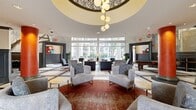 This sophisticated 2,050-square-foot lobby and common area occupies a single floor and is designed to impress from the moment of entry. The grand entrance corridor features floor-to-ceiling glass walls on both sides, flooding the space with natural light, and is crowned by a striking multicolored blown-glass chandelier suspended from soaring heights above white-and-gray patterned tile flooring.

The main lobby area is anchored by bold, terra-cotta-red cylindrical columns with decorative bases, which define zones throughout the open-plan space. A dramatic curved mezzanine balcony with ornate wrought-iron railings overlooks the seating area below, which features a large-scale clock wall mural as a focal art installation. Curated lounge areas are furnished with dark leather sofas, gray upholstered armchairs, and sculptural accent pieces, including a distinctive white-and-gold pod side table.

A secondary lounge zone benefits from an impressive wall of floor-to-ceiling windows offering direct views of an outdoor pool and courtyard, creating a seamless indoor-outdoor connection. A recessed ceiling detail with a globe pendant cluster light fixture adds architectural interest overhead. Transitional corridors feature elegant wainscoting in slate-blue tones with black trim and classic tile patterning. Throughout, the space balances contemporary furnishings with refined architectural detailing.