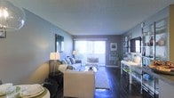 This single-floor space centers around a well-appointed living area that balances comfort and style. Dark hardwood floors run throughout, providing a rich contrast against the soft, neutral grey walls and textured ceiling. The room is generously sized, offering ample space for multiple seating arrangements, including a plush sofa accented with decorative pillows and a coordinating armchair positioned near the back of the room.

A large sliding glass door dominates the far wall, flooding the interior with abundant natural light and offering a glimpse of a private outdoor area beyond. Vertical blinds frame the door, allowing for adjustable light control. The layout is open and functional, with a defined seating zone anchored by a soft area rug and a low-profile coffee table.

On one side of the room, a dedicated media area features a wall-mounted flat-screen television atop a sleek console, complemented by open shelving. Tasteful wall art and a decorative mirror add character to the space, while carefully placed floor lamps enhance the warm, inviting ambiance. Overall, the living area presents a clean, contemporary aesthetic suited for both relaxing and entertaining.