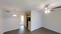 This single-floor residence offers a thoughtfully arranged layout that promotes a natural flow between living spaces. The main living area features rich, dark hardwood-style flooring that extends throughout, creating a warm and cohesive aesthetic. A ceiling fan with a light fixture keeps the space comfortable and well-lit, while a pendant light adds a subtle decorative touch near the dining area.

The kitchen is positioned just off the main living space, partially enclosed by a dividing half-wall that maintains an open feel while defining the cooking area. Dark appliances and upper cabinetry are visible within the kitchen, giving it a functional and streamlined appearance. A window in the kitchen area allows natural light to filter in.

Beyond the main living area, a passage leads to an additional room featuring carpeted flooring and a window that brings in natural light, offering a softer, more subdued atmosphere compared to the rest of the home. The neutral wall tones throughout the residence provide a clean, versatile backdrop suitable for a variety of interior styles. The overall ambiance is practical and inviting, with a straightforward single-level design that makes the space easy to navigate and live in.