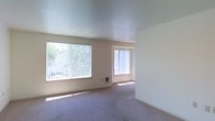 This single-floor space features an open living area finished with wall-to-wall carpeting in a neutral gray-purple tone, contributing to a quiet and comfortable atmosphere. The room is illuminated by two generously sized windows fitted with vertical blinds, allowing natural light to filter in and brighten the interior throughout the day. The windows face outward toward green foliage, providing a pleasant outdoor connection. The walls are painted in a soft, warm beige, complementing the light-toned textured ceiling above. The layout is straightforward and open, offering flexible furniture arrangement options. A wall outlet is centrally positioned along the main wall, adding practical utility to the space. The clean lines, neutral palette, and abundance of natural light give the area a simple, functional character well-suited for everyday living.