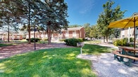 Located at 4839 W College Ave in Greendale, Wisconsin, this single-story brick building is part of a well-established residential community characterized by its mature, park-like grounds. The exterior showcases classic brick construction with large windows that provide ample natural light throughout. The building's façade is complemented by neatly trimmed hedges and maintained landscaping that lines the entryways.

The surrounding grounds are a defining feature of this property. Towering mature trees — including prominent evergreens and broad deciduous varieties — frame the central courtyard, creating shaded walkways and open lawn areas throughout. Mulched beds are interspersed with green grass, giving the outdoor space a well-tended, park-like atmosphere. Paved pathways wind through the grounds, connecting the buildings and providing accessible routes for residents.

A central courtyard area includes a picnic table with an umbrella and decorative lamp posts, offering a comfortable outdoor gathering space. Bicycle racks are also visible along the walkways, indicating a pedestrian-friendly environment. The open layout between the buildings enhances the sense of space and fosters an inviting community atmosphere under the open Wisconsin sky.