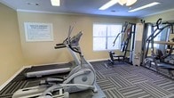 This single-floor fitness center offers a practical and functional workout environment for residents. The space is outfitted with essential exercise equipment, including a treadmill, an elliptical cross-trainer, and a multi-station cable weight machine, providing options for both cardiovascular and strength training workouts. The room features a striped, low-pile carpet that adds a degree of comfort underfoot while maintaining a clean, structured aesthetic. Warm beige walls and crown molding give the space a polished, residential feel. Natural light filters in through a window fitted with horizontal blinds, complementing the overhead recessed lighting to keep the space well-lit throughout the day. A posted rules board on the wall helps ensure shared use of the facility remains organized and respectful for all users. The overall ambiance is straightforward and purpose-driven, prioritizing accessibility and functionality. This fitness center serves as a convenient on-site amenity, allowing residents to maintain an active lifestyle without leaving the property.