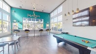 Located at 20080 West Dixie Highway in Miami, FL, this dynamic single-floor clubroom serves as a lively social hub designed for recreation and community gathering. The space immediately impresses with its soaring double-height ceilings and an expansive wall of floor-to-ceiling windows that flood the interior with abundant South Florida natural light, framing views of the surrounding palm-lined outdoor areas and pool.

The design aesthetic is bold and contemporary, anchored by a striking teal accent wall adorned with decorative circular wall art and colorful framed artwork throughout. Stylish pendant lighting clusters hang at varying heights, adding warmth and visual interest to the open layout. Polished stone-look tile flooring runs throughout, lending a clean, modern foundation to the space.

A full-size billiards table serves as a central recreational focal point, complemented by a bar-height counter with yellow-accented swivel stools — ideal for socializing. Multiple dining and work tables with mixed seating provide flexible space for gatherings or casual meetings. A wall-mounted flat-screen television adds to the entertainment options.

The kitchenette area is well-appointed with stainless steel appliances including a refrigerator, microwave, and under-counter drawers, paired with white quartz countertops. Large sliding glass doors open directly to an outdoor patio, creating a seamless indoor-outdoor connection perfect for Miami's year-round climate.