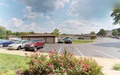 This single-story property offers a practical and well-organized exterior layout designed to accommodate residents with ease and convenience. The grounds feature a clearly marked parking area with ample spaces, complemented by a row of detached garage units with brown overhead doors — providing secure, covered storage for vehicles. Additional covered carport structures extend the parking options further across the property.

The landscaping adds a welcoming touch to the exterior, with vibrant rose bushes in full bloom lining the walkways and softening the edges of the parking area. Mature trees dot the background, contributing to a green and pleasant setting. The surrounding terrain is gently rolling, offering open views toward the horizon under a wide, partly cloudy sky.

The building itself is constructed in a low-profile, single-story style with neutral-toned siding and a simple, functional roofline consistent across the garage and main structures. The paved driveways and lot are well-maintained, ensuring clear circulation throughout the property. Overall, the space presents a tidy, suburban character with a strong emphasis on practical amenities including dedicated parking, garage access, and thoughtfully maintained outdoor areas.