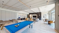This expansive single-floor amenity space offers a sophisticated blend of recreation, wellness, and social gathering areas, all finished with contemporary design elements throughout. The clubroom serves as the centerpiece, featuring a vibrant blue billiard table, a ping-pong table, and a dedicated workspace with built-in desks and curated wall art. A comfortable lounge area with modern upholstered seating surrounds a large wall-mounted TV set against a striking dark shiplap accent wall, complemented by beamed ceilings and light wood-style flooring. Floor-to-ceiling windows and glass doors flood the lounge with natural light and provide views of the outdoor surroundings. The catering kitchen is outfitted with a large stainless steel island with bar seating, pendant lighting, open shelving, patterned tile backsplash, and full-size stainless steel appliances. A sliding barn door leads to the fitness wing, which includes a fully mirrored weight and cardio room stocked with a wide range of equipment, as well as a separate group fitness and cycling studio with large windows, ceiling fans, and ample open floor space for classes and training. Throughout the space, curated artwork, recessed lighting, and cohesive neutral tones create a polished, welcoming atmosphere.