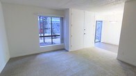 This single-floor space offers a straightforward, functional layout with a clean and neutral aesthetic throughout. The main living area features wall-to-wall carpeting in a neutral tone, contributing to a quiet and comfortable atmosphere underfoot. White walls provide a bright, blank canvas that allows for flexible use of the space.

Natural light enters through large windows, which frame views of an exterior stairwell and the surrounding building structure. A secondary windowed area is visible toward the rear of the space, further enhancing the sense of openness and connectivity to the outdoors. The exterior views reflect a multi-unit building environment with structured outdoor staircases.

The ceiling features a textured finish, adding a subtle visual element to the otherwise minimalist interior. A solid white door provides access to an adjacent room or exterior area, maintaining the clean, uncluttered character of the space. The overall layout is open and adaptable, with no major visual obstructions dividing the primary living zone. The combination of neutral finishes, carpeted flooring, and ample window placement makes this a practical and versatile space suitable for a variety of uses.