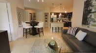 This single-floor residence showcases a thoughtfully designed open-concept layout that seamlessly connects the living, dining, and kitchen areas into one cohesive and inviting space. The living area features a sleek gray leather sofa, a distinctive triangular glass coffee table, and a patterned area rug layered over light-toned tile flooring that runs throughout the space, contributing to a bright and airy atmosphere.

The dining area is anchored by a rich dark-wood pub-style table paired with white upholstered high-back chairs, offering a stylish contrast that adds visual depth. Adjacent to the dining space, the kitchen is outfitted with dark cabinetry, stone countertops, and stainless steel appliances, including a refrigerator and microwave. A kitchen island with bar seating provides additional casual dining and prep space, illuminated by pendant lighting overhead.

Track lighting and recessed fixtures throughout the open living area ensure the space remains well-lit, enhancing the warm, neutral color palette of the walls and furnishings. A hallway visible from the main living area leads to additional rooms, with built-in shelving offering functional storage. The overall ambiance is modern and polished, combining dark wood accents with light surfaces for an elegant and practical everyday living environment.