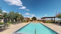 Located at 1103 Bernard St in Denton, Texas, this outdoor amenity area is defined by its classic brick architecture and well-maintained landscaping. The entrance is framed by substantial stone and brick columns supporting a covered entryway, with wrought iron gates providing a secure and welcoming access point. Brick-paved walkways and neatly trimmed hedgerows contribute to a polished, manicured exterior aesthetic, while vintage-style lantern lighting adds a refined touch to the surroundings.

The centerpiece of the space is a generously sized outdoor swimming pool with crystal-clear water, bordered by a wide concrete deck that offers ample room for relaxation. A pair of wooden pergolas flank the pool area, providing shaded retreats, while multiple lounge chairs and patio seating are arranged throughout the deck for comfort. Lush mature trees line the perimeter, offering natural shade and a sense of privacy. A raised water feature is visible at the far end of the pool, adding a decorative architectural element. The open-sky setting allows for abundant natural light, creating a bright and inviting outdoor environment ideal for leisure and recreation year-round.