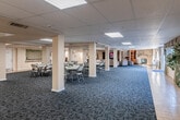 This single-floor community space is thoughtfully designed to accommodate a variety of social and recreational activities. The main hall is expansive, featuring a drop-panel ceiling with recessed fluorescent lighting and patterned blue-gray carpeting that extends throughout most of the space. Cream-colored support columns are evenly distributed across the hall, defining the open layout without obstructing the sense of space. Round tables with folding chairs are arranged throughout, making the area well-suited for events, meetings, or communal dining.

A dedicated lounge area offers a more relaxed setting, furnished with comfortable gray sofas, wooden entertainment cabinetry, and a large flat-screen television. A stone accent wall anchors this corner of the space, adding warmth and texture to the overall aesthetic.

Adjacent to the lounge, a game room features a full-size pool table beneath skylights that bring in natural light, accompanied by wingback chairs for casual seating. Framed artwork lines the walls, contributing a welcoming atmosphere.

The entry corridor is bright and welcoming, with tile flooring, draped windows allowing natural light, and decorative plants near the entrance. Bulletin boards and framed notices line the hallway, reflecting an active and engaged community environment.