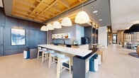 Located at 12004 Inspiration St in Reston, VA, this expansive single-floor amenity space delivers a polished, contemporary environment defined by thoughtful architectural details and high-end finishes throughout. Floor-to-ceiling glass curtain walls flood the space with abundant natural light and frame sweeping views of the surrounding Reston skyline, creating a bright and open atmosphere at every turn.

The grand lobby serves as a striking focal point, featuring large-format stone tile flooring, textured stone-clad accent walls, and curated abstract artwork. Dramatic oversized pendant light fixtures anchor distinct seating zones, where sculptural lounge chairs and plush sofas invite gathering and relaxation. A dedicated media lounge area includes a wall-mounted display screen framed by a bold dark panel surround.

The kitchen and dining area showcases a coffered wood ceiling with integrated lighting, providing warmth and visual texture. A long white-topped island with bar seating and a contrasting dark base serves as the centerpiece, complemented by open shelving, full cabinetry, and professional appliances. Intimate booth-style seating alcoves with warm wall sconces offer a more private setting for smaller gatherings. Throughout the space, the interplay of natural wood, matte stone, and soft neutral tones establishes a cohesive, sophisticated ambiance.