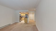 This single-floor unit offers a well-organized layout that maximizes functionality across its living spaces. The open-concept design connects the living area and kitchen seamlessly, with neutral-toned carpet throughout the main living and bedroom areas providing a warm, cohesive feel. The kitchen is a standout feature, equipped with stainless steel appliances including a refrigerator, electric range, and over-the-range microwave. Light maple cabinetry extends to the upper walls, offering generous storage, while dark granite-style countertops add a polished contrast. A peninsula with matching countertop serves as a natural divider between the kitchen and the living area, offering additional workspace. Recessed lighting throughout the kitchen ensures a bright, functional environment, complemented by tile flooring. The bedroom is a quiet, carpeted retreat featuring a wall-mounted air conditioning unit and baseboard heating, providing year-round climate control. A full bathroom includes a tiled tub-shower combo, light tile flooring, and a toilet with overhead storage — clean and straightforward in design. A hallway connects the main areas, with a door providing access to an additional entry or utility space. The overall layout is practical and efficient, suited for comfortable everyday living.