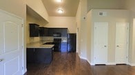This single-floor space makes a strong visual impression through its combination of high ceilings and an open layout that enhances the sense of volume throughout. The kitchen serves as a central feature, outfitted with dark espresso-toned cabinetry that extends nearly to the ceiling, providing generous storage capacity. Granite countertops in a light, variegated tone offer a striking contrast against the dark cabinetry, while a full suite of black appliances — including a refrigerator, range, microwave, and dishwasher — maintains a cohesive, contemporary aesthetic. A peninsula counter extends into the space, offering additional prep area and the potential for casual seating. Rich, dark hardwood flooring runs throughout, anchoring the space with warmth and durability. The soaring ceiling height creates an airy, open ambiance, drawing the eye upward and amplifying the overall sense of space. Neutral warm-toned walls provide a versatile backdrop throughout. A white-paneled door is visible along the adjacent wall, suggesting access to additional rooms or storage. Overhead flush-mount lighting provides consistent illumination throughout the kitchen and adjoining areas.