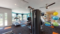 This fitness center in Jackson, Tennessee, is a single-floor facility designed to support a comprehensive range of workout routines. The interior is defined by vibrant teal accent walls contrasted against white surfaces, creating an energetic atmosphere. The flooring consists of multi-toned carpet tiles in navy, orange, and blue, providing both visual interest and functional cushioning throughout the space.

The facility is organized into distinct workout zones. The cardio area is anchored by treadmills and elliptical machines positioned along a curved, floor-to-ceiling window wall that floods the space with natural light and offers outdoor views of the surrounding greenery. Ceiling fans provide additional ventilation throughout.

The strength training section houses a multi-station cable machine, a free-weight bench press setup with Olympic barbells and weight plates, and additional resistance equipment. Full-length mirrors along one wall allow users to monitor their form during exercises.

A dedicated functional training area features upright and recumbent stationary bikes, spin bikes, and a well-stocked accessories wall displaying medicine balls, resistance bands, stability balls in multiple sizes, and foam rollers. Multiple wall-mounted flat-screen televisions are positioned throughout the space for entertainment. Recessed lighting provides consistent illumination across all workout zones.