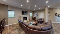 This single-floor entertainment space is thoughtfully designed to accommodate socializing and recreation. The main lounge area anchors the layout, featuring a generous sectional leather sofa and matching recliner arranged around a rustic wood coffee table. A wall-mounted flat-screen TV sits above a sleek electric fireplace framed by a white mantel, creating a welcoming focal point. Recessed lighting throughout keeps the space well-lit, while neutral taupe walls and wood-look flooring lend a warm, cohesive aesthetic.

Adjacent to the lounge, a dedicated game area houses a full-size pool table beneath a pendant light, offering an engaging recreational option. A decorative column and open sightlines connect this zone seamlessly to the rest of the floor.

The kitchen and bar area is a standout feature, centered on a substantial curved bar with stacked stone cladding and a granite countertop. Industrial-style globe pendant lights hang above, illuminating the space. Bar-height seating lines the perimeter, and the kitchen behind it includes dark cabinetry, a refrigerator, microwave, and sink. A large whiteboard on a nearby wall adds a functional, event-ready touch to this well-appointed communal space.
