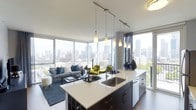 This contemporary urban residence showcases a thoughtfully designed open-concept layout that seamlessly integrates the kitchen, dining, and living areas into a single, cohesive living space. Floor-to-ceiling windows wrap around the space, flooding the interior with abundant natural light while framing dramatic, panoramic views of the surrounding city skyline — a defining feature that gives the space an elevated, cosmopolitan character.

The kitchen is anchored by a large central island finished with a crisp white countertop and contrasting dark cabinetry, offering generous prep space and a built-in sink. A trio of sleek pendant lights hang above the island, providing both functional task lighting and a striking visual element. The cabinetry's dark tones create a bold contrast against the bright, airy surroundings.

The living area flows naturally from the kitchen, benefiting from the same expansive window views. The space accommodates a comfortable seating arrangement with ample room to move freely, all set atop rich hardwood-style flooring that adds warmth to the modern aesthetic. The neutral palette throughout is accented with pops of color, creating an inviting yet refined atmosphere. Overall, the space strikes a balance between sleek, modern design and livable comfort with remarkable urban vistas.