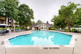 Located at Holliswood Court in Charlotte, NC, this outdoor amenity area serves as a welcoming community hub centered around a generously sized swimming pool with clear, blue water. The pool features a geometric, multi-angled shape with clearly marked depth indicators and no-diving designations, catering to a wide range of recreational use. Surrounding the pool is a broad concrete deck furnished with blue lounge chairs and side tables, providing ample space for relaxation and outdoor gatherings. The focal point of the area is a charming single-story clubhouse constructed with stone masonry and accented by a prominent arched window with white trim, French-style glass doors, and a pitched roof with a chimney. The structure exudes a classic, cottage-like character that complements the natural surroundings. Mature deciduous trees frame the entire space, offering shade and a lush, park-like atmosphere throughout the area. A wrought iron perimeter fence encloses the pool area, ensuring safety while maintaining visual openness to the landscaped grounds. The multi-story residential buildings visible in the background, each featuring balconies, reinforce the sense of community this amenity space is designed to serve.