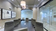 This single-floor conference room exudes a polished, professional ambiance designed to facilitate focused meetings and collaborative discussions. At the heart of the space sits a substantial dark oval conference table surrounded by high-backed leather executive chairs in a warm taupe tone, offering both comfort and a refined aesthetic.

The room's design details elevate it above a standard meeting space. Crown molding frames the ceiling, lending a classic architectural touch, while a striking chandelier — featuring a drum shade paired with an ornate candelabra-style fixture — serves as a central focal point and provides warm, layered lighting alongside recessed ceiling lights.

A standout architectural feature is the arched glass window centered on the far wall, which floods the room with diffused natural light and creates an open, airy feel. A full-height glass partition wall to one side maintains visual connectivity with adjacent spaces while preserving acoustic privacy.

The neutral beige walls and dark hardwood flooring create a sophisticated contrast, complemented by a framed artwork piece on the left wall. The overall atmosphere is formal yet inviting, making it well-suited for professional presentations, executive meetings, or client consultations.
