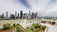 This rooftop space offers a striking urban retreat set against the full panorama of the Downtown Los Angeles skyline. The space is defined by its expansive, open-air layout, where clear glass panel perimeter railings preserve unobstructed sightlines to the city's iconic high-rises from every vantage point.

A key design feature is the integrated green roof system, which incorporates generously sized raised planting beds filled with drought-tolerant succulents, agaves, and low-growing groundcovers. A separate area of the rooftop introduces a grove of young trees planted in deep, rust-toned raised planters, alongside vibrant red-foliaged shrubs, lending a naturalistic, layered quality to the hardscape.

The surface underfoot consists of clean, light-toned concrete pavers that complement the neutral, contemporary palette of the surrounding parapet walls. Access to the rooftop is provided through glazed entry doors, which allow views of the skyline and gardens from within. Supporting utilities and safety equipment are neatly integrated into the perimeter walls. The overall ambiance is one of sophisticated, eco-conscious urban design — a functional yet serene outdoor environment that balances horticultural greenery with dramatic, uninterrupted views of the Los Angeles cityscape.