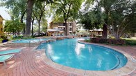 Located on Bellaire Ranch Drive in Fort Worth, Texas, this outdoor amenity complex offers a resort-inspired environment designed for relaxation and recreation. At the heart of the space is a generously sized freeform swimming pool with a connected spa, both framed by classic red brick pavers and surrounded by mature shade trees that provide a naturally cooled atmosphere. Adjacent to the pool, a covered pergola with a wood-slatted roof and ceiling fans shelters a fully equipped outdoor kitchen featuring stainless steel grills, granite countertops, and stone masonry bases — ideal for outdoor entertaining. A spacious wooden deck serves as a central gathering area, furnished with comfortable seating and anchored by a striking stacked-stone outdoor fireplace, creating a welcoming focal point for cooler evenings. The grounds also include a putting green set within a well-manicured lawn, adding a recreational dimension to the outdoor experience. Brick-paved walkways wind through lush, mature landscaping and connect the various amenity areas. Stone and brick architectural detailing throughout the complex ties together a cohesive Texas Hill Country aesthetic, complemented by wrought iron fencing and gated access points that provide both security and visual appeal.