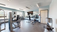 Located at 5085 Main St in Trumbull, Connecticut, this well-appointed fitness center offers a comprehensive workout facility across a single open floor. The space is designed with functionality and comfort in mind, featuring light blue-gray walls, durable speckled rubber flooring, and modern linear LED lighting throughout.

The main workout area is generously sized and organized into distinct training zones. A dedicated cardio section houses multiple elliptical trainers and treadmills, positioned near large windows that allow ample natural light and outdoor views of the surrounding property. A strength training zone features a multi-station cable machine and bench press setup, while a free weights area provides a full rack of dumbbells, weight benches, and stability balls for versatile training options.

Full-length mirrors line one wall of the main exercise floor, visually expanding the space and providing exercisers with proper form feedback. A wall-mounted flat-screen television adds convenience for entertainment during workouts.

The facility is accessible via glass-paneled entry doors and includes a transition area near the entrance, offering a clean and organized point of entry. The thoughtful layout ensures sufficient open floor space between equipment stations, allowing for safe and comfortable movement throughout the room.