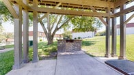 Located at 4960 Edmondson Pike in Nashville, Tennessee, this outdoor pavilion serves as a communal gathering space within a residential community. The structure features a wood-framed gabled roof supported by sturdy wooden posts, providing ample shade over a concrete patio floor. A metal picnic table with bench seating sits at the center of the covered area, offering a comfortable spot for outdoor dining and socializing. Adjacent to the pavilion stands a built-in stone masonry grill, constructed with stacked stone detail and designed for outdoor cooking and barbecuing. The pavilion opens on all sides, encouraging airflow and offering unobstructed views of the surrounding green lawn, mature trees, and the neighboring residential buildings. The lush, well-maintained grounds extend outward from the structure, creating an inviting outdoor environment. Additional seating and utility amenities are visible nearby, further supporting the space's role as a shared outdoor hub. The open-air design, combined with the natural canopy of surrounding trees, makes this pavilion a practical and pleasant outdoor amenity for the community it serves.