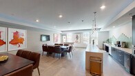 This contemporary single-floor amenity space is thoughtfully designed to accommodate both casual gatherings and structured events. The open-plan layout is anchored by large-format porcelain tile flooring that flows seamlessly throughout, lending a cohesive and polished aesthetic.

The fully equipped kitchen is a standout feature, boasting dark espresso cabinetry with brushed gold hardware, a striking geometric green hexagonal tile backsplash, stainless steel appliances including a built-in refrigerator and wall oven, and an expansive white quartz island topped by elegant globe pendant lights. The kitchen transitions naturally into the main lounge area, where a lengthy white quartz countertop bar provides additional workspace and casual seating.

The dining zone accommodates a large dark wood table surrounded by rich brown leather chairs, positioned beneath recessed lighting and adjacent to a wall-mounted flat-screen television. Textured grasscloth-style wall panels add warmth and dimension to the space. Curated contemporary artwork punctuates the walls throughout.

A dedicated game area features a foosball table and additional seating, bathed in natural light from multiple windows with horizontal blinds. A glass partition wall elegantly separates zones while maintaining visual openness. Lush tropical plants and bold abstract art complete the refined yet inviting atmosphere of this well-appointed communal space.